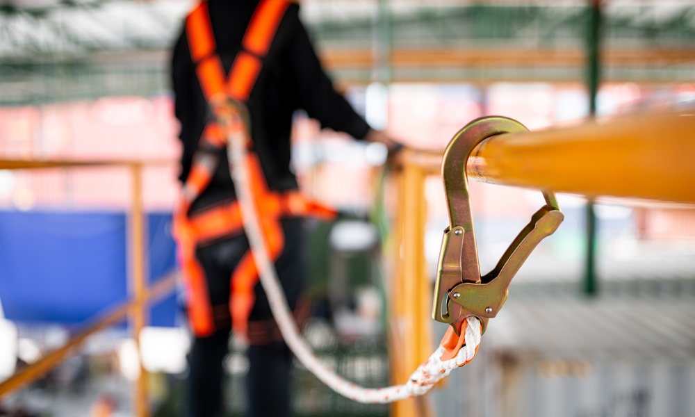A person wearing an orange safety harness connected by a white rope to a metal hook attached to a yellow railing.