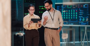 Two coworkers reviewing information on a tablet in a server room with illuminated equipment and digital displays.