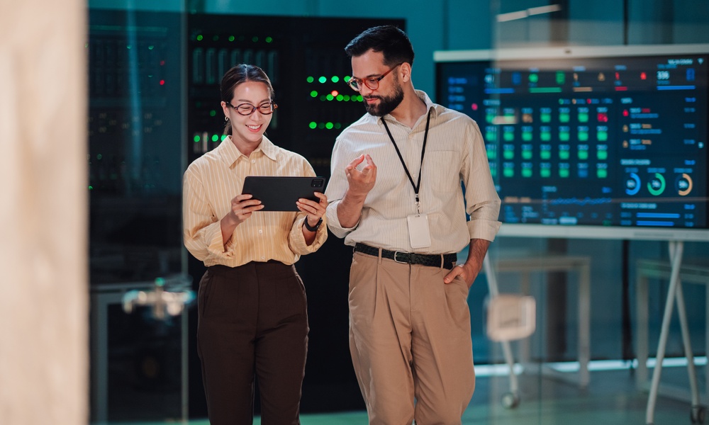 Two coworkers reviewing information on a tablet in a server room with illuminated equipment and digital displays.