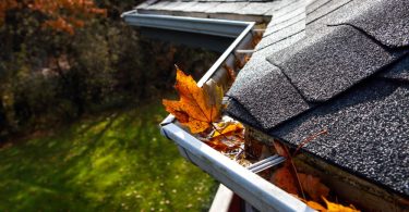 Close-up of a residential roof with asphalt shingles and rain gutters clogged with fallen autumn leaves.