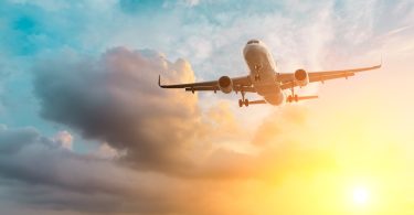 A commercial passenger airplane flying overhead with its landing gear extended against dark clouds and a golden sunset sky.