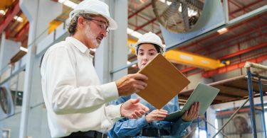 A man in a white shirt and hard hat holding a clipboard beside a woman in a blue jumpsuit and hard hat using a tablet.