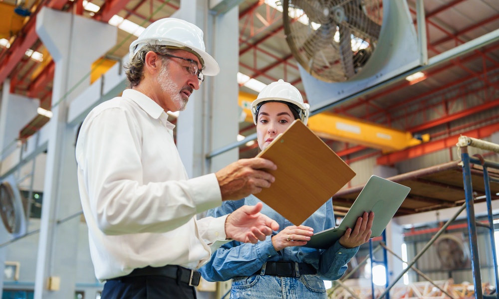 A man in a white shirt and hard hat holding a clipboard beside a woman in a blue jumpsuit and hard hat using a tablet.