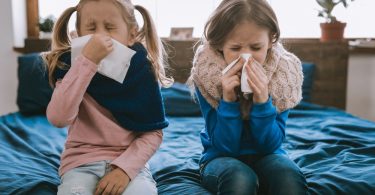 Two young girls sit on the edge of a bed as they blow their noses from being sick. They both wear scarves.
