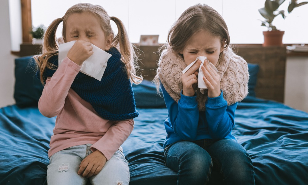 Two young girls sit on the edge of a bed as they blow their noses from being sick. They both wear scarves.