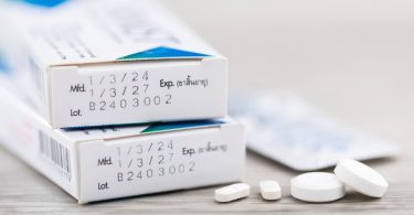 A few white medication tablets sit on the table next to the pharmacy boxes. The box shows the dates for expiry and use.