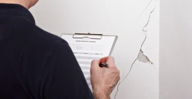 A man in a dark shirt writing on a clipboard beside a white wall with a long crack and an exposed patch of plaster.