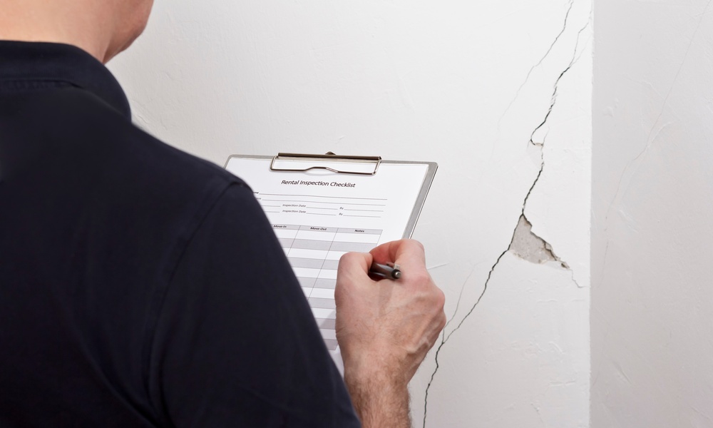A man in a dark shirt writing on a clipboard beside a white wall with a long crack and an exposed patch of plaster.