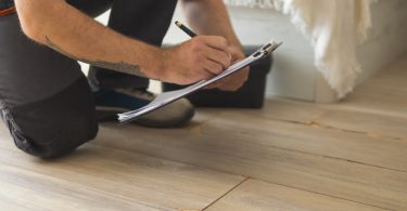 A person kneeling on a light wood floor while writing on a clipboard beside a cracked and lifted laminate plank.