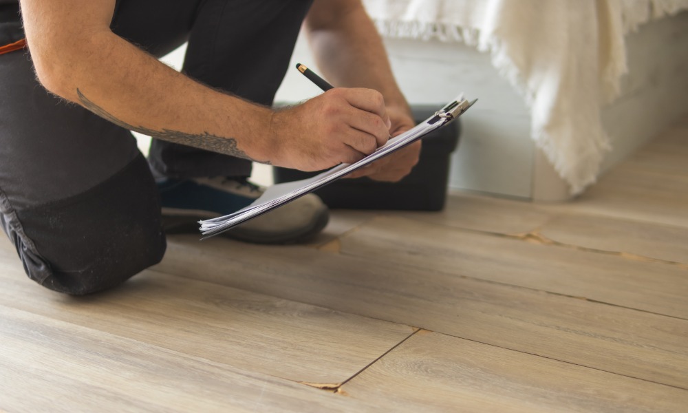 A person kneeling on a light wood floor while writing on a clipboard beside a cracked and lifted laminate plank.