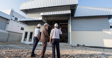 Three people wearing white hard hats standing outside a large industrial building, with one gesturing toward its entrance.