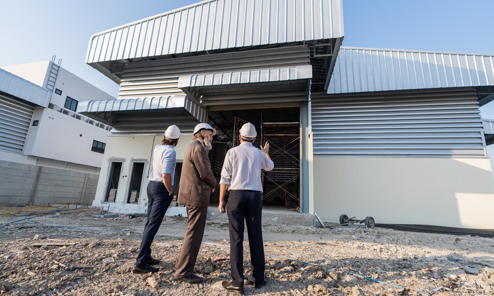 Three people wearing white hard hats standing outside a large industrial building, with one gesturing toward its entrance.