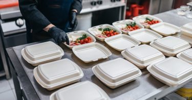 A food service worker preparing meals in open takeout containers on a stainless steel kitchen counter.