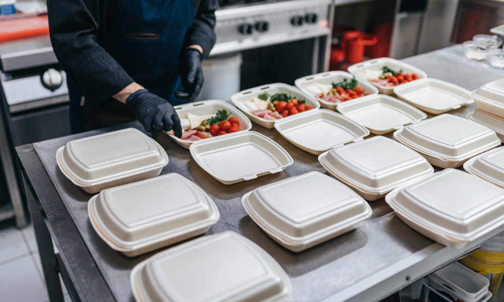 A food service worker preparing meals in open takeout containers on a stainless steel kitchen counter.
