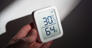 A close-up of a homeowner's hand holding an indoor thermostat with temperature and humidity on the display.