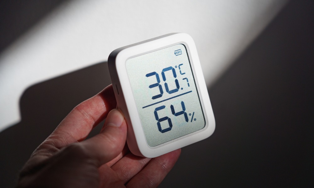 A close-up of a homeowner's hand holding an indoor thermostat with temperature and humidity on the display.