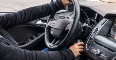 A person in a long-sleeved shirt holding a steering wheel with one hand and turning the ignition key with the other.