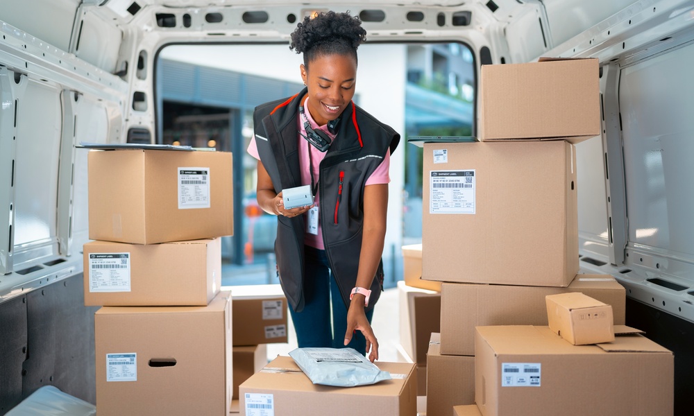 A woman wearing a black vest over a pink shirt handling a small parcel and a scanner inside a van filled with boxes.