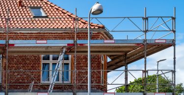 Scaffolding surrounds an older brick building with a red tile roof and white-framed windows during exterior renovation work.