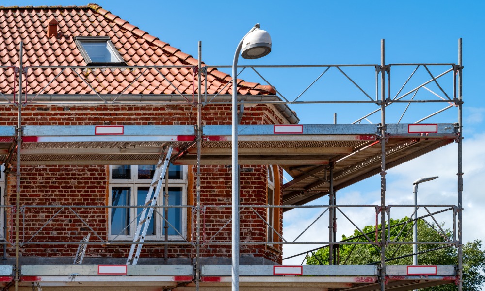 Scaffolding surrounds an older brick building with a red tile roof and white-framed windows during exterior renovation work.