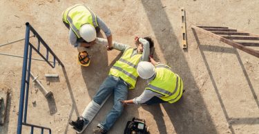 A construction worker on the ground with his leg in a cast. Two other construction workers are tending to his injuries.