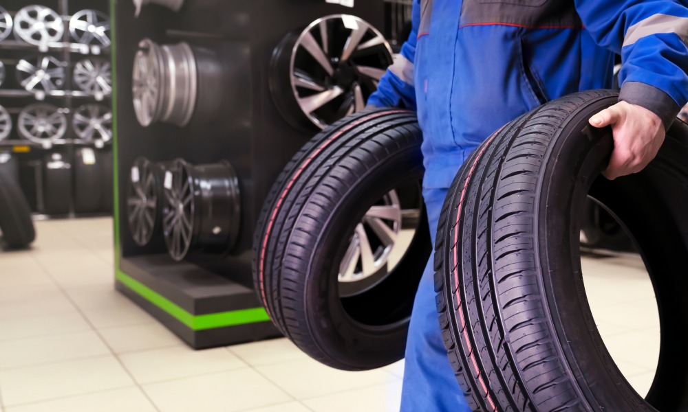 A tire shop technician holding two new vehicle tires, with a display of alloy wheels and rims in the background.