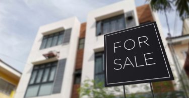 A "For Sale" sign is displayed in front of a multi-story residential building with brick accents and large windows.