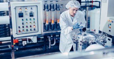 A woman in white clothing is standing in an industrial setting looking at a machine. She has a clipboard.