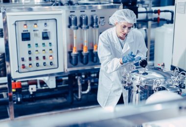 A woman in white clothing is standing in an industrial setting looking at a machine. She has a clipboard.