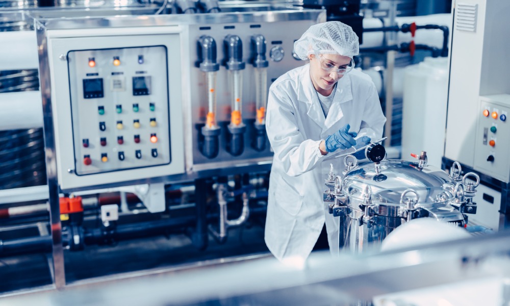 A woman in white clothing is standing in an industrial setting looking at a machine. She has a clipboard.