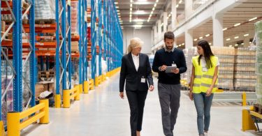 A man and two women are walking through a warehouse facility. The man is holding a clipboard.