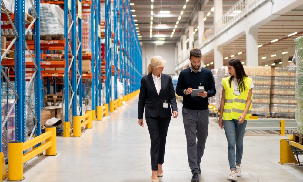 A man and two women are walking through a warehouse facility. The man is holding a clipboard.