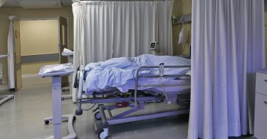 A hospital room with a moving bed covered with light blue sheets, surrounded by fabric curtains under bright light.