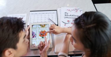 Two people reviewing floral arrangements on a tablet, with wedding planning notebooks and a calendar on the table.