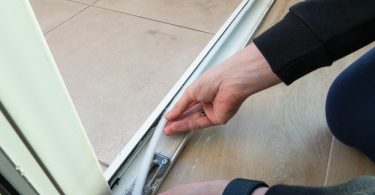 A person wearing a long-sleeve black shirt is applying white sealing tape on the bottom track of a residential door.