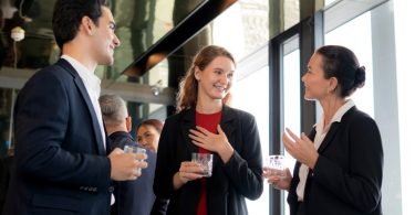 Two women wearing suits, talking to each other at an event with a man standing next to them in a dark suit.
