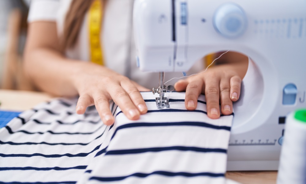 A close-up of two hands holding a white piece of fabric with blue stripes under the needle of a sewing machine.