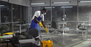 A custodian wearing a face mask using a mop to clean the floor of an office. There are desks set up around him.