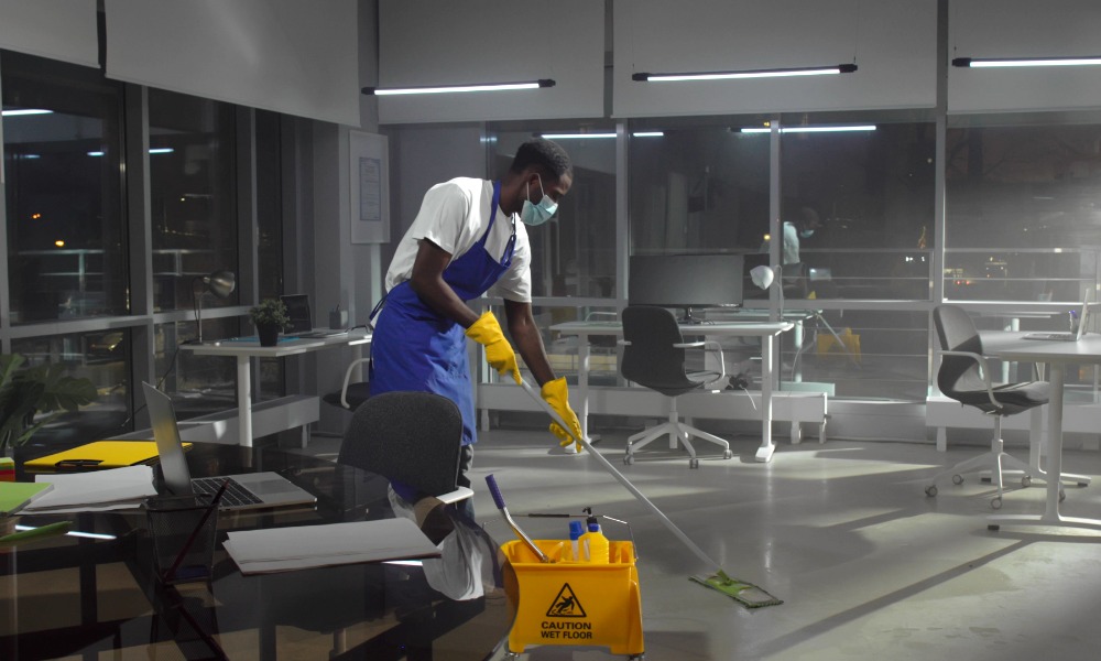 A custodian wearing a face mask using a mop to clean the floor of an office. There are desks set up around him.