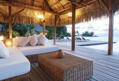 Luxury beachfront cabana with white cushioned sofas, wicker furniture, and a thatched roof overlooking an infinity pool.