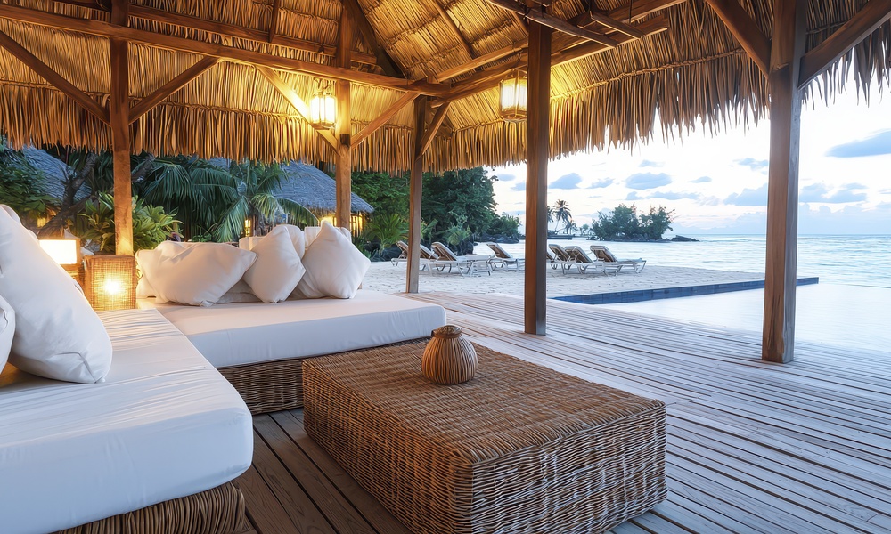 Luxury beachfront cabana with white cushioned sofas, wicker furniture, and a thatched roof overlooking an infinity pool.