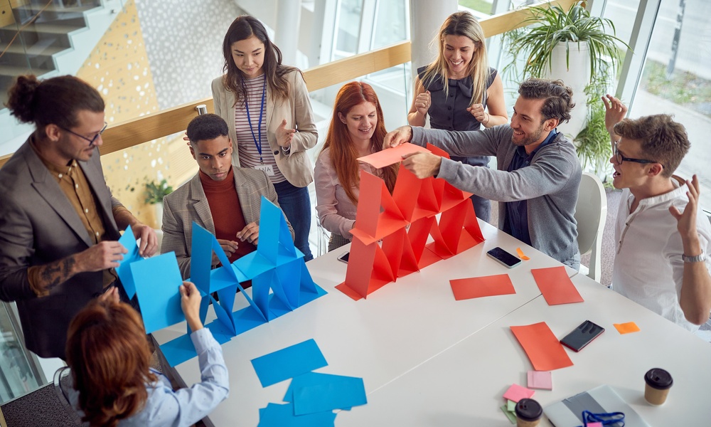 Employees construct blue and red paper structures around a conference table.