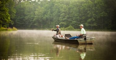 Two men sit in a small fishing boat on a lake. The man in the front of the boat is casting a fishing line.