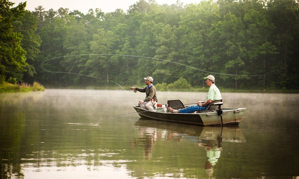 Two men sit in a small fishing boat on a lake. The man in the front of the boat is casting a fishing line.