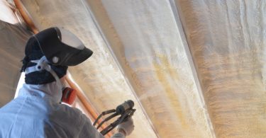 A worker wearing a white uniform and a large face mask using a spray foam gun to cover an angled roof in a room.