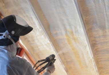 A worker wearing a white uniform and a large face mask using a spray foam gun to cover an angled roof in a room.
