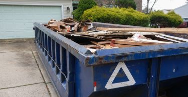 A blue dumpster is parked on a residential driveway. There is a house in the background of the shot.