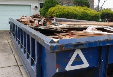 A blue dumpster is parked on a residential driveway. There is a house in the background of the shot.