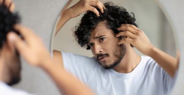 A man looking in a mirror as he pulls his hairline back to inspect it. He is looking for signs of hair loss.
