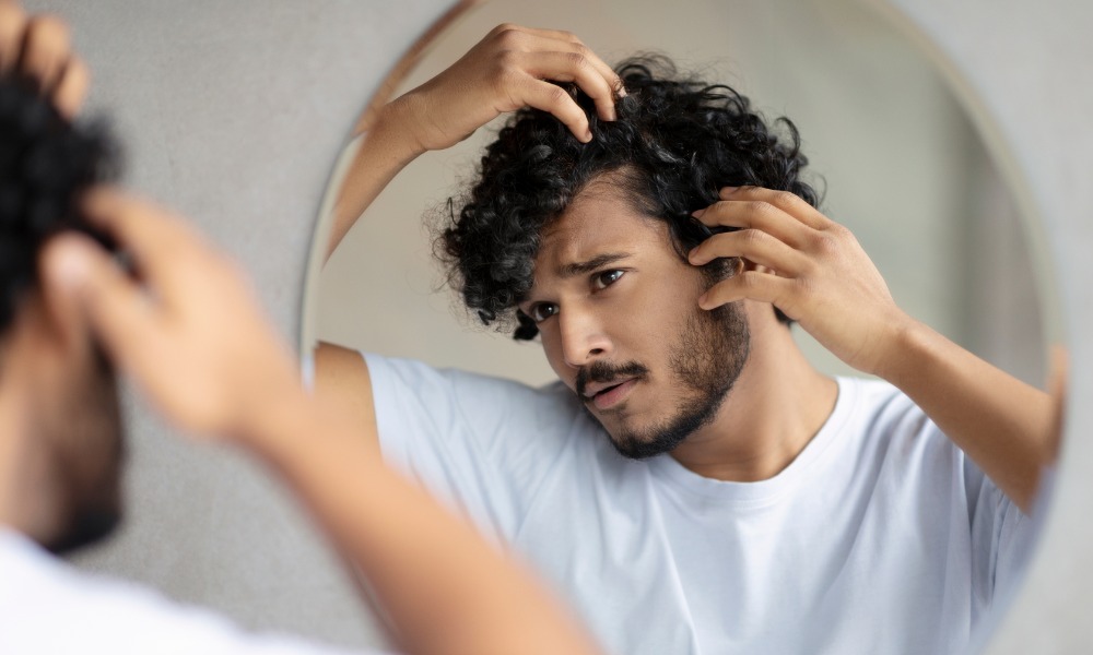 A man looking in a mirror as he pulls his hairline back to inspect it. He is looking for signs of hair loss.